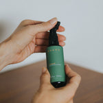


Hand holding a green spray bottle with a white label on a wooden surface.